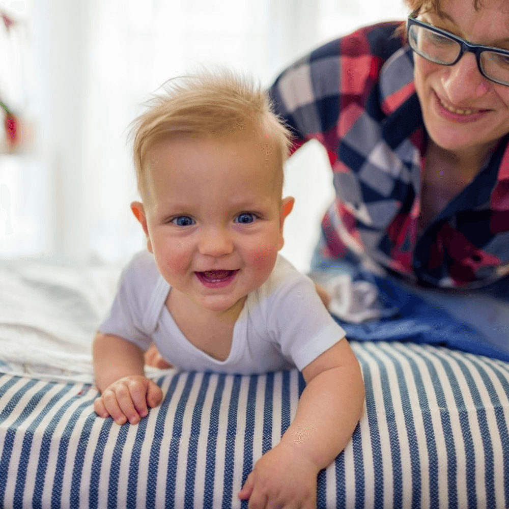 Smiling baby laying on the top of a bed next to a caretaker 