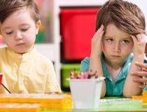 Boy in green t shirt with hands on his head looking frustrated, he is sitting next to another boy doing an activity at a desk