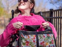 Lady is smiling and sitting in a wheelchair, wearing a pink jumper and holding a bag