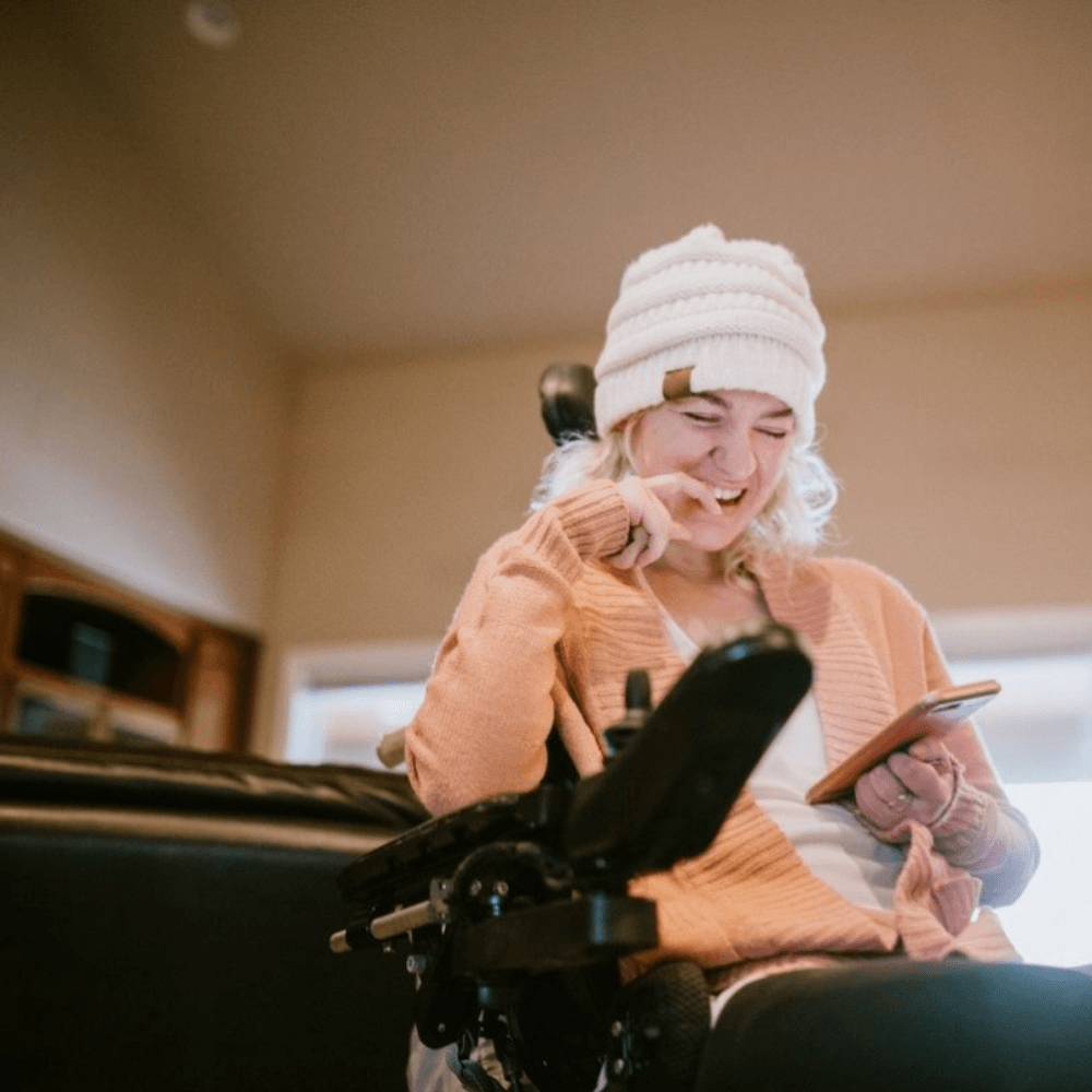 Lady wearing a white beanie, smiling in wheelchair, and looking at her phone