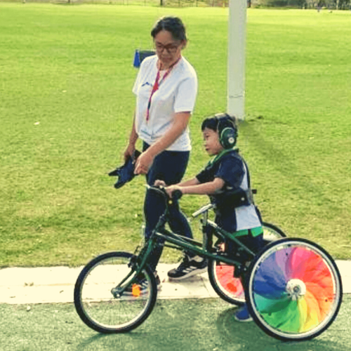 A mum is next to her son writing a bike with colourful wheels.