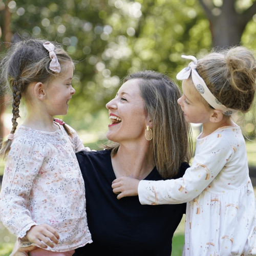 A mum is smiling at her two daughters wearing white. They are in a park.